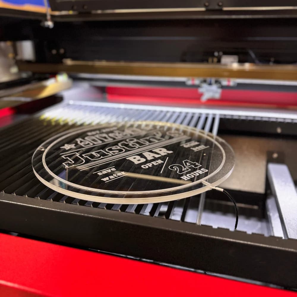 A personalized circular acrylic sign for "Justin's Bar" being precision-etched in a laser engraver, featuring a "24 Hours Open" design for a custom LED man cave display.