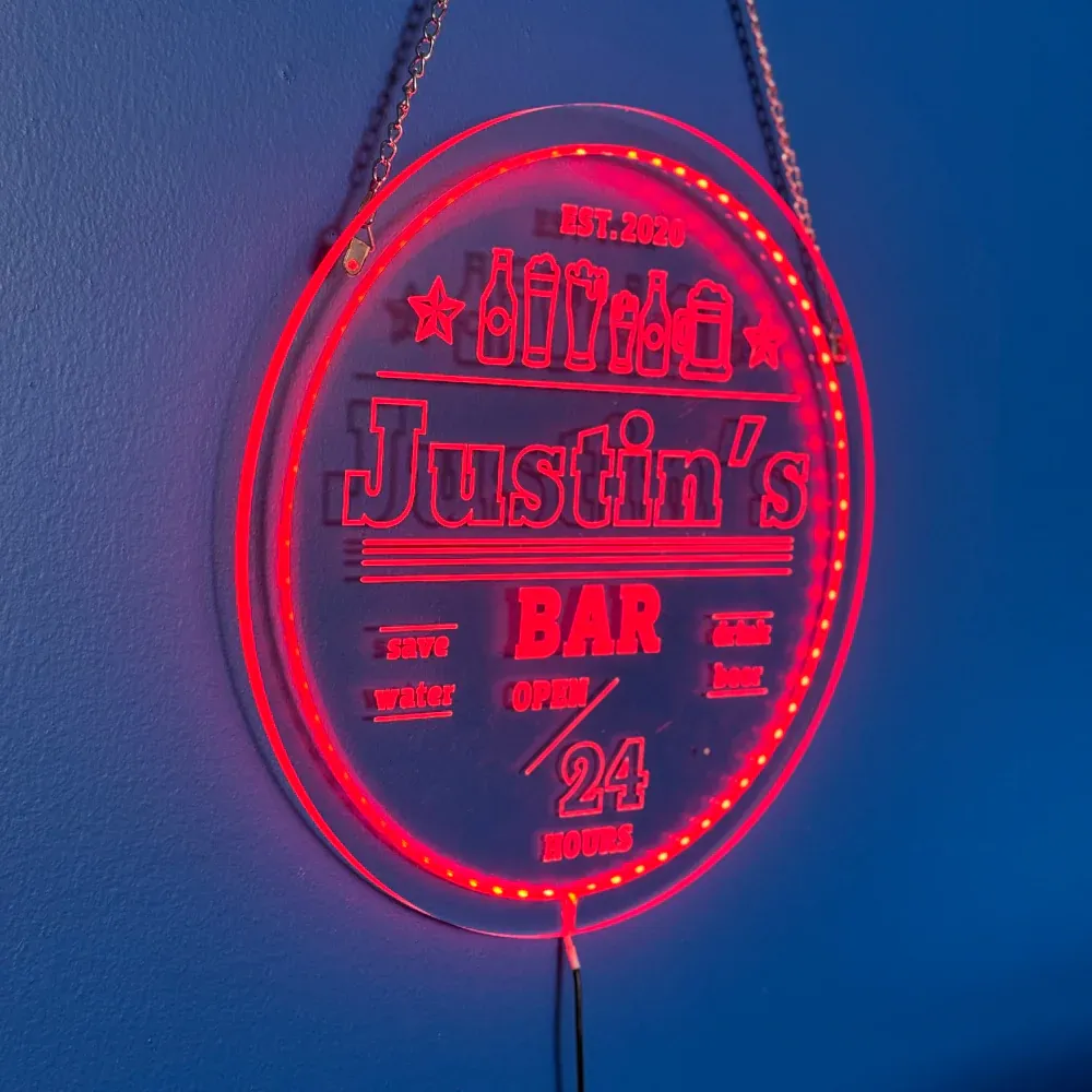 An oblique close-up view of the finished personalized circular LED sign for "Justin's Bar," glowing brightly with red light on a textured blue wall. The sign is suspended by chains and features detailed etchings of beer bottles, stars, and the text "EST. 2020," "BAR," "save water drink beer," and "OPEN 24 HOURS."